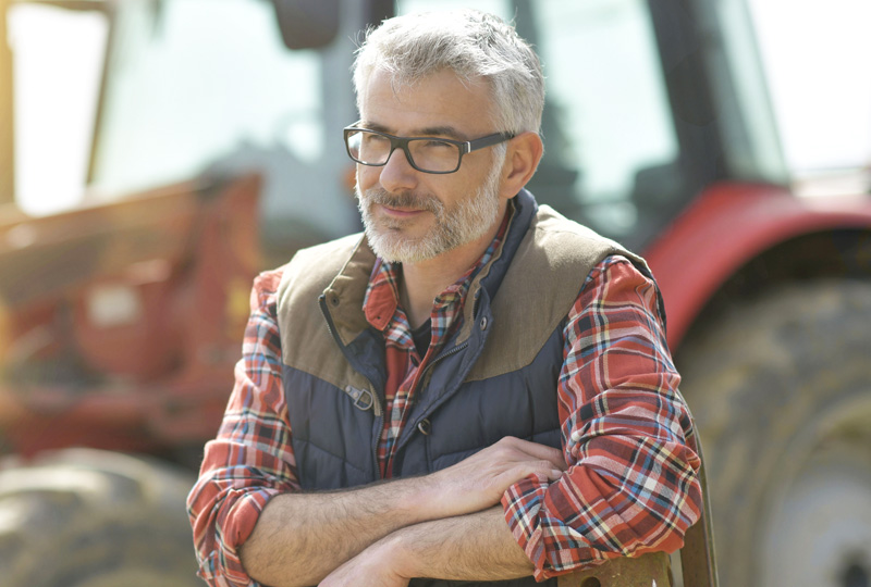 mature man wearing glasses while standing in front of a tractor outdoors